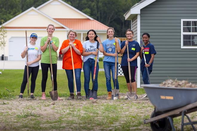 Seven Community Engagement Scholars holding shovels and rakes in a yard.